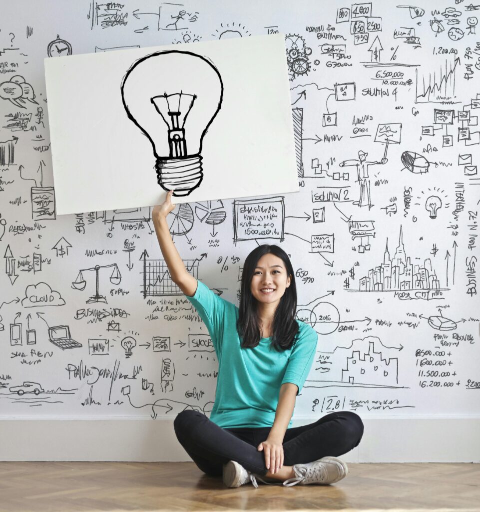 pexels-photo-3758105-3758105 Young woman holds a light bulb drawing against a creative brainstorming wall.