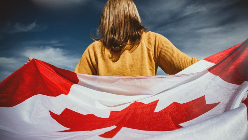 Your Pathway to Healthcare Licensing in Canada Rear view of a woman in a yellow sweater holding a large Canadian flag against a blue sky.