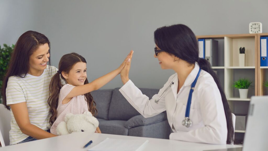 qualified healthcare professionals A smiling female doctor giving a high-five to a young girl sitting with her mother in a medical office.