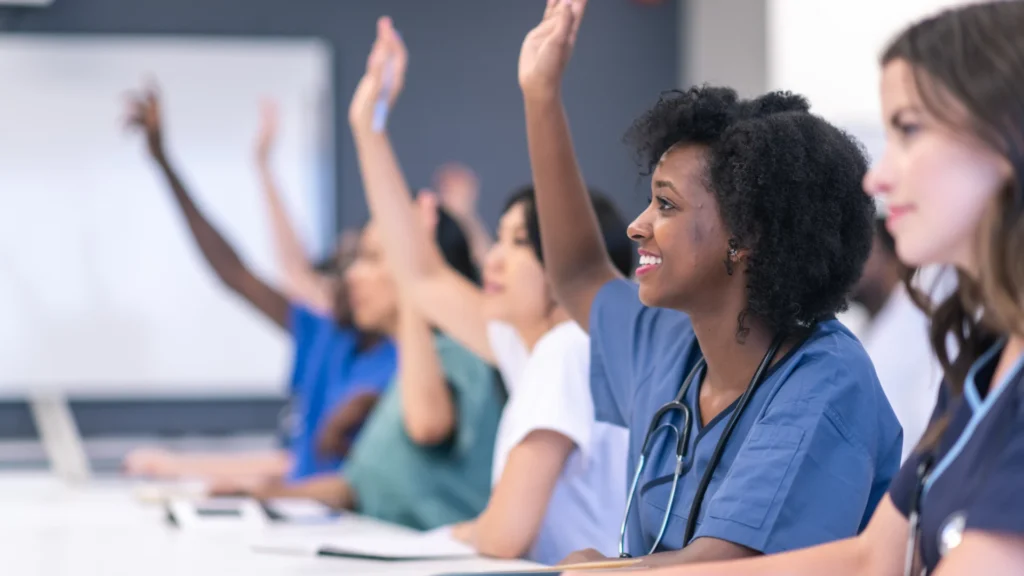 A diverse group of healthcare professionals in blue and green scrubs raising their hands to participate in a medical seminar classroom.