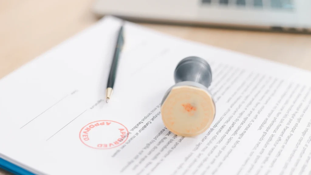 Close-up of an official document on a desk featuring a red "APPROVED" rubber stamp next to a grey stamp and a pen.