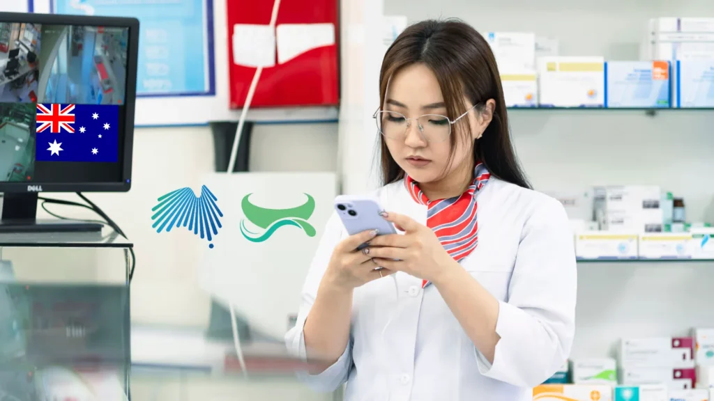 A female pharmacist in a white lab coat using a smartphone in a pharmacy, with a monitor displaying the Australian flag and healthcare regulatory logos.