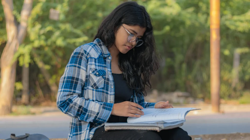 A young woman with long dark hair and glasses sitting outdoors, focused on studying a large open textbook on her lap.