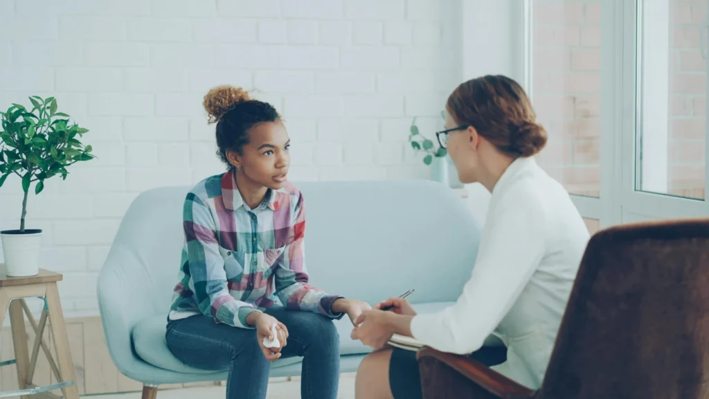 A young woman with curly hair and a plaid shirt sits on a sofa, in discussion with a professional consultant in a white blazer who is holding a notebook.