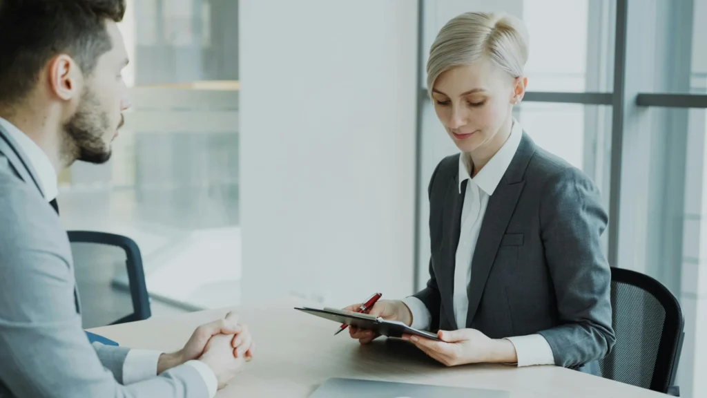 A male job applicant with a beard is being interviewed by a focused female interviewer in a grey suit holding a clipboard in a modern office.
