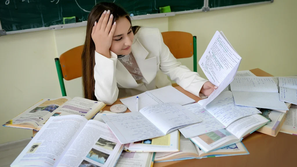 A young woman in a white blazer studying at a desk covered in textbooks and notebooks, preparing for a Prometric or Pearson VUE medical licensing exam.