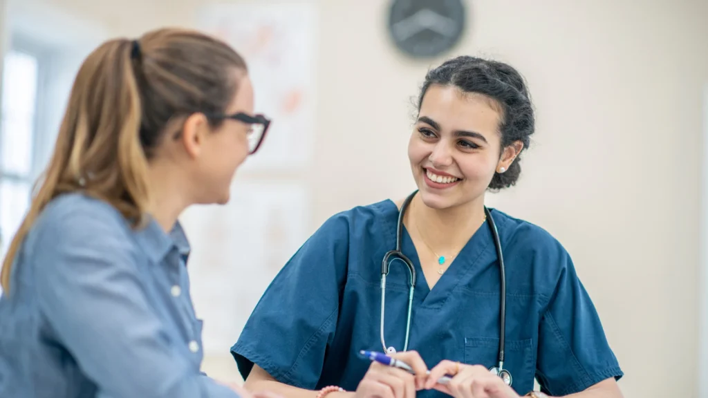 A professional female healthcare provider in navy blue scrubs and a stethoscope smiling in a bright, modern clinic.