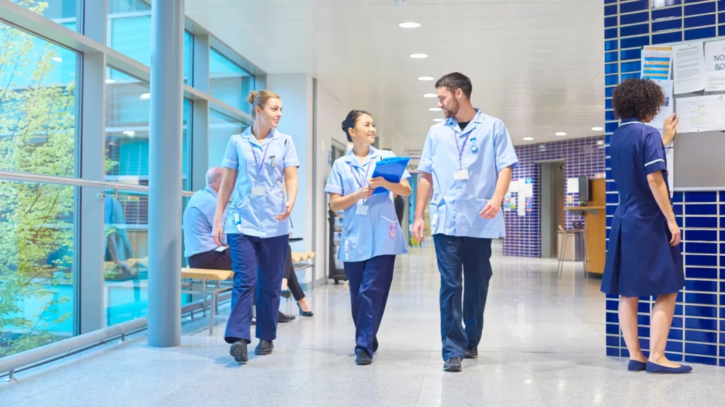 A diverse team of three nurses in light blue uniforms walking and talking in a bright, modern hospital corridor.